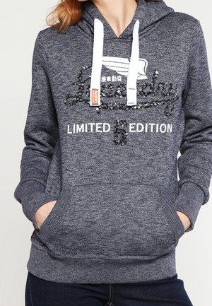 Persoon die een grijze hoodie draagt met witte aantrekkoorden, een geborduurd grafisch ontwerp en de tekst "LIMITED EDITION", handen in de voorzak.