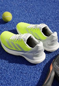 Neon yellow and gray sports shoes on a blue tennis court next to a tennis ball and a black and orange racket handle.