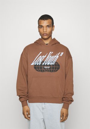 Brun hoodie med frontlomme, med hvid og sort tekstgrafik. Blødt stof, afslappet pasform og hætte med snoretræk. Lyseblå jeans.