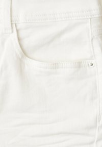 Gros plan d'un tissu en denim blanc montrant la partie supérieure d'une poche avant avec des coutures et un rivet métallique.