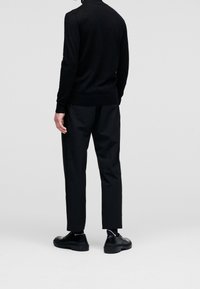 Černý pletený svetr s těsnými manžetami a lemem, spárovaný s černými kalhotami. Černá obuv s texturovanou podrážkou dokládá celkový vzhled.