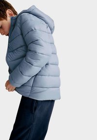 Ljusblå vadderad jacka med huva, med horisontell vaddering och dragkedja. Bärs av ett barn, kombinerad med mörka byxor.