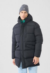 Hombre vistiendo una chaqueta acolchada negra con capucha, gorro gris, capa interior turquesa y jeans azul claro contra un fondo liso claro.