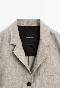 Veste gris clair en tissu texturé, col à revers cranté, et un bouton noir. L'étiquette à l'intérieur indique "Massimo Dutti", précisant la marque et l'origine.