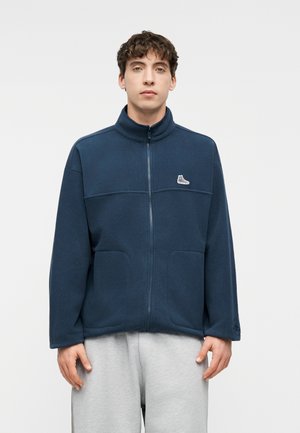 Mladý muž s krátkymi kučeravými tmavými vlasmi, oblečený v námornícky modrej mikine na zips a svetlosivých nohaviciach, stojaci na jednofarebnom pozadí.