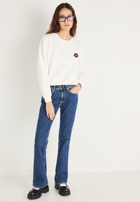 Felpa bianca con grafica di labbra rosse, abbinata a jeans a zampa di elefante blu e scarpe platform nere. Modella con occhiali, in piedi contro uno sfondo neutro.