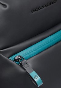 Sac noir avec une fermeture éclair turquoise, une tirette texturée avec logo, et un matériau lisse. Design simple aux lignes épurées.