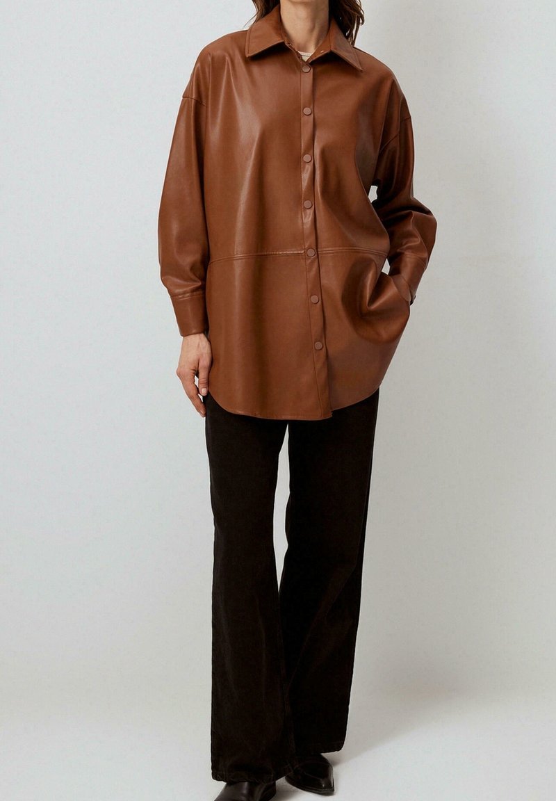 Chemise surdimensionnée en cuir marron avec boutonnage devant, col pointu et manches longues, associée à un pantalon noir à jambes larges. Texture lisse.