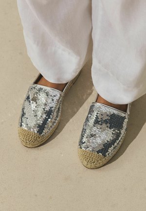 Feet wearing silver sequin espadrilles with braided jute toes, paired with loose white linen pants on a beige floor.