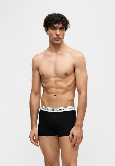 Νέος άνδρας με σγουρά μαλλιά φοράει μαύρα boxer briefs με λευκή ζώνη Polo Ralph Lauren, στέκεται μπροστά σε απλό φωτεινό φόντο.
