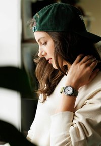 Noor naine, kes kannab rohelise mütsi tagurpidi ja valget sviitrit, puudutab oma kaela, käe peal on rohelise rihmaga kell, millel on valge taust.