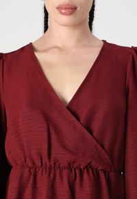 Rochie burgundă, cu decolteu adânc în V, dintr-o țesătură texturată și talie strânsă. Mânecile tip puff adaugă un detaliu structural designului.