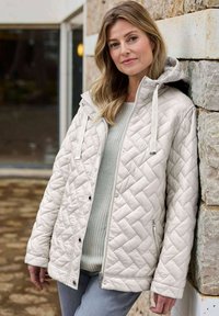 Hellbeige Steppjacke mit Kapuze, diagonalem Steppmuster, Frontreißverschluss und Seitentaschen, getragen über einem leichten Pullover.