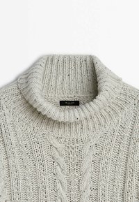 Gros plan sur un pull à col roulé en maille épaisse couleur crème, avec un motif torsadé et un col côtelé.
