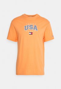 Oranžové bavlněné tričko s kulatým výstřihem, na kterém je výrazně napsáno "TOMMY USA JEANS" vzorovanými písmeny a malou vlajkovou nálepkou na spodní straně.