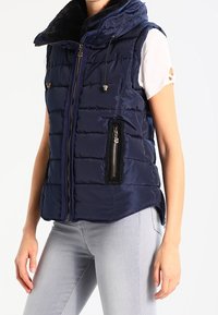 Femme portant une veste sans manches matelassée bleu marine avec des poches zippées par-dessus une chemise blanche à manches courtes et un jean gris clair.