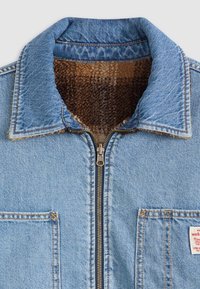 Chaqueta de denim azul claro con cuello puntiagudo, cierre de cremallera y forro de felpa marrón. Cuenta con un bolsillo en el pecho y detalles de costura en naranja.