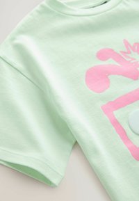 Tricou verde deschis din bumbac moale, cu decolteu rotund. Are un design grafic mare roz și un accent circular în centru.