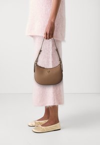 Brown leather handbag with a curved design and gold hardware, held by a person wearing a pink fuzzy top and pink skirt, paired with beige flats.
