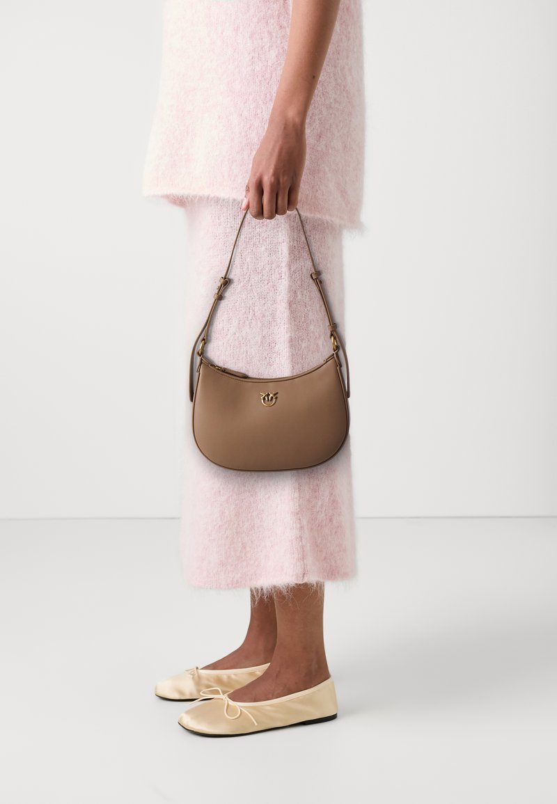 Brown leather handbag with a curved design and gold hardware, held by a person wearing a pink fuzzy top and pink skirt, paired with beige flats.