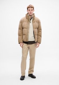 Beige pufferjakke med høj krave, lynlås og kviltet struktur, parret med et cremefarvet indre lag og beige bukser. Sorte sko.