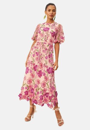 Vrouw draagt een roze midi-jurk met pofmouwen en geborduurde paarse en roze bloemen, gecombineerd met sandalen met hoge hak en bandjes.