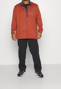 Giacca in pile rossa con zip frontale, dettagli grigi e una tasca sul petto a sinistra. Indossata con pantaloni neri e scarpe da ginnastica nere.