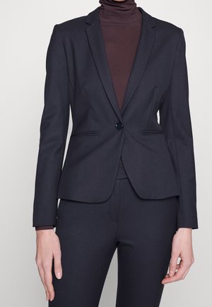 Mujer con blazer negro ajustado con un botón sobre un jersey de cuello alto marrón oscuro y pantalones negros a juego sobre un fondo liso.