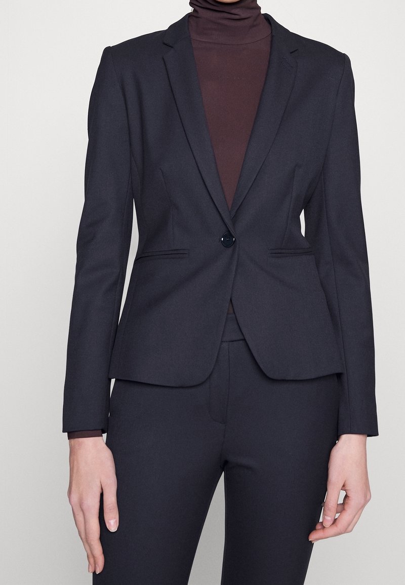 Femme portant un blazer noir cintré avec un bouton sur un col roulé marron foncé et un pantalon noir assorti sur un fond uni.