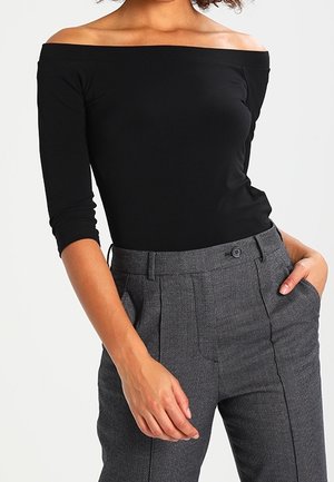Vrouw draagt een nauwsluitende zwarte off-shoulder top met driekwart mouwen en hooggetailleerde grijze, op maat gemaakte broek met zakken.