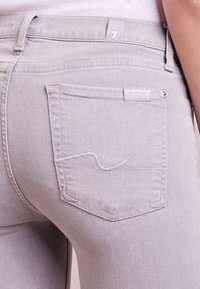 Jeans skinny de color gris claro con un único bolsillo trasero, con costuras blancas sutiles y una etiqueta cerca de la cintura. Textura de denim suave.