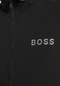 Musta vetoketjullinen takki, joka on valmistettu teksturoidusta kankaasta. Takissa on korkea kaulus ja heijastava painettu logo "BOSS" rinnan kohdalla.