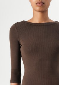 Brown ribbed long-sleeve top with a boat neckline, fitted shape, and textured fabric. Features subtle stitching along the neckline.