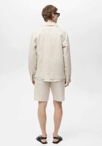 Svetlo bež lanena srajca z ujemajočimi se kratkimi hlačami, z sproščenim krojem, kratkimi rokavi in črnimi sandali, prikazana s hrbtne strani.