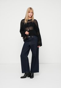 Schwarzer Strickpullover mit weiten Ärmeln, kombiniert mit hochtaillierten, dunkelblauen, weit geschnittenen Jeans. Die Schuhe sind schwarze Stiefeletten.