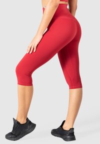 Leggings capri rouges en tissu extensible, dotés d'une taille haute et d'un design épuré. Associés à des chaussures de sport noires.