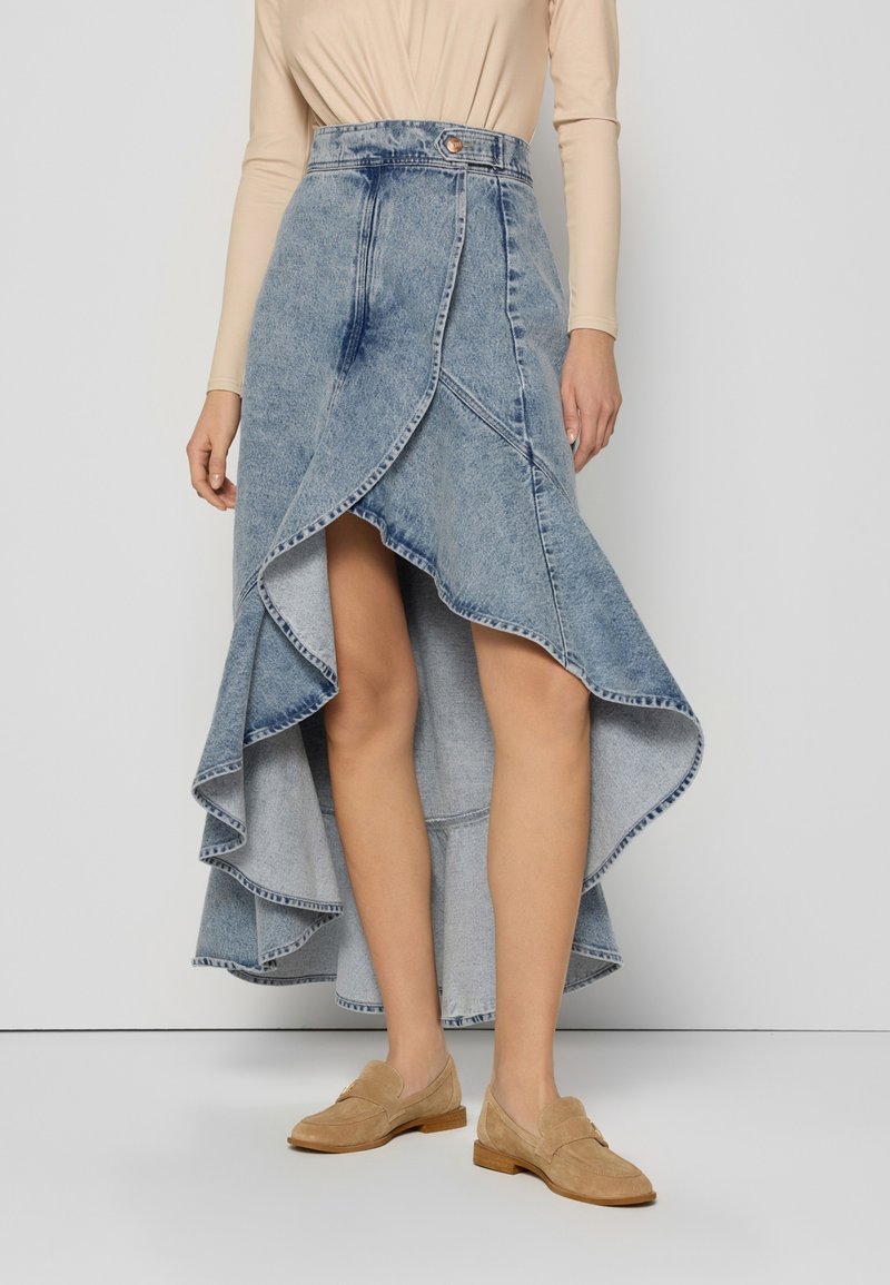 Denim high-low rok met een flare zoom, rauwe randen en een lichte blauwe wassing. Gecombineerd met tan instapschoenen, die een gelaagd ontwerp tonen.