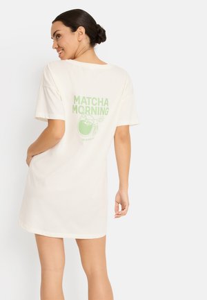 Femme aux cheveux foncés attachés en chignon portant une robe t-shirt blanche oversize avec le texte vert « Matcha Morning » et un motif de thé dans le dos.