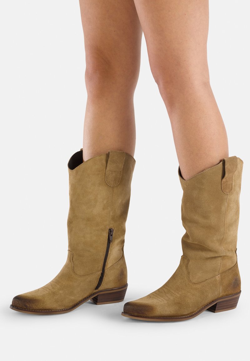 Stivali cowboy alti in camoscio color tan con punta affusolata, zip laterale e bordo superiore angolato. Presentano cuciture testurizzate sulla punta e un tacco basso.