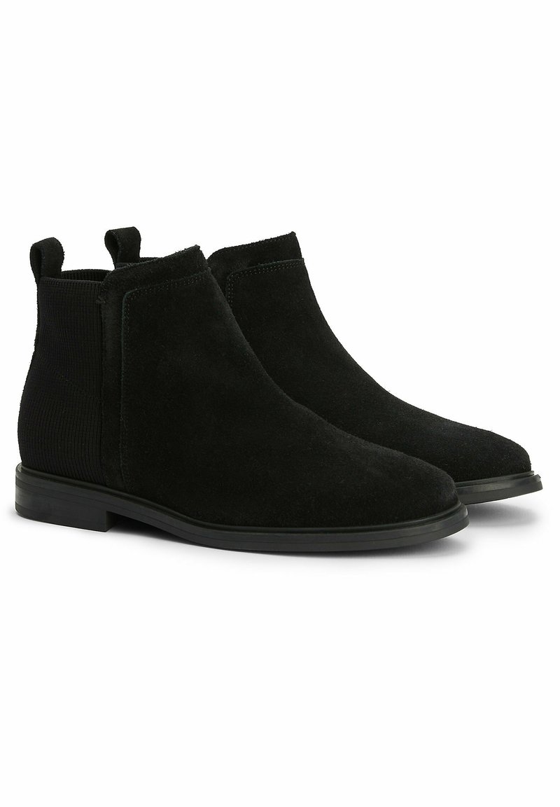 Paire de bottines Chelsea en daim noir avec panneaux élastiques sur les côtés et languettes à tirer, semelles plates noires, photographiées sur fond blanc.