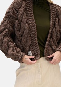 Gruby sweter rozpinany w ciemnobrązowym kolorze z warkoczowymi wzorami, otwartym przodem i prążkowanymi brzegami. Nosi się go na ciemnozielony golf. Widoczna metka.