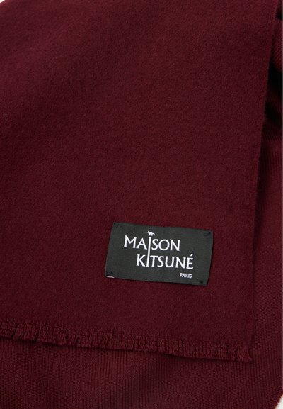 Μπούργκουντι μάλλινο κασκόλ με φραγκοσυταγωγές άκρες. Διαθέτει μια μαύρη ετικέτα με λευκό κείμενο "MAISON KITSUNÉ PARIS" στην κάτω γωνία. Μαλακή υφή.