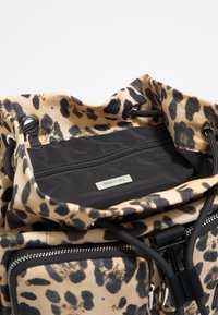 Torba na sznurki z tkaniny w panterkę z czarnymi akcentami, z wewnętrzną kieszenią na zamek i metalową plakietką marki. Teksturowana powierzchnia z różnorodnymi plamkami.
