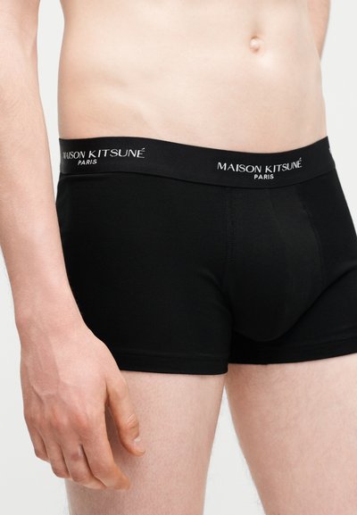 Αρσενικός κορμός και άνω πόδια με μαύρα εσώρουχα boxer με την ένδειξη «Maison Kitsuné Paris» στη ζώνη, πάνω σε απλό φόντο.