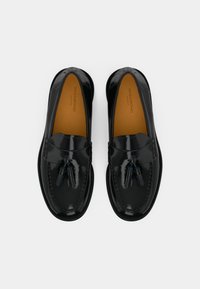 Black patent leather loafers with a rounded toe, featuring a decorative tassel accent and contrasting tan insole. Smooth surface texture.