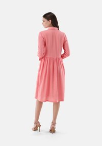 Rosa, langärmliges Kleid mit Kragen, geraffter Taillenpartie und knielangem Saum. Der Stoff wirkt leicht und leicht strukturiert.