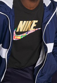 Svart bomullströja med en gyllene "NIKE"-logotyp och en färgglad Swoosh-design som är gjord av olika nationalflaggor. Marinblå jacka under.