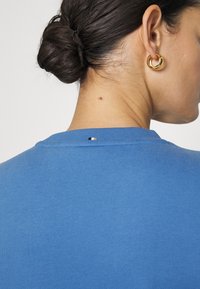 Blue cotton t-shirt featuring a round neck, smooth texture, and a small black and white accent on the back. Ear accessory is a gold hoop.