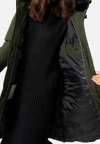 Olivengrøn puffet jakke med lynlås og hætte, med en sort ribstrikket sweater under. Indvendigt foret med skinnende sort stof, inkluderer en lomme.