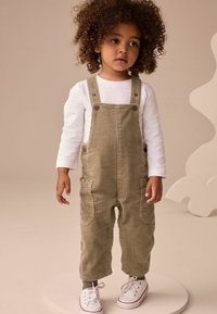 Salopette en velours côtelé olive avec des rayures horizontales, dotée de deux poches avant, de boutons-pression en métal et de poignets retroussés, associée à une chemise blanche à manches longues.